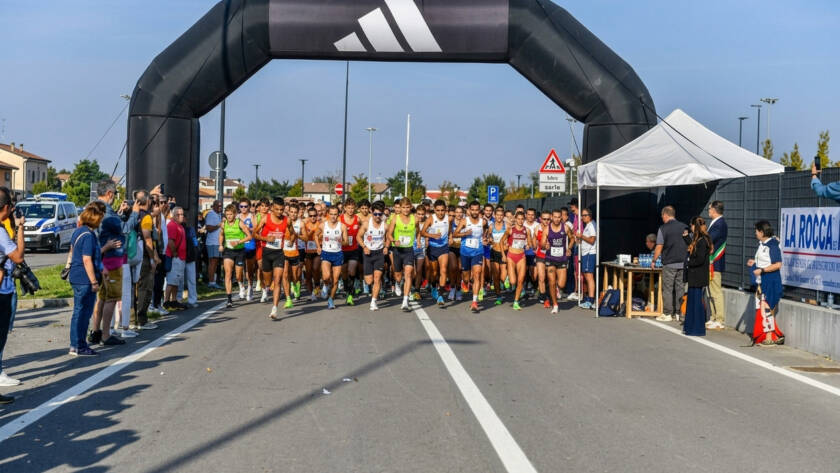 Gruppo numeroso di podisti in tenuta sportiva al momento della partenza della gara Nova Run, mentre attraversano la linea sotto un grande arco gonfiabile nero targato Adidas. Sulla destra sono presenti un gazebo bianco, transenne con spettatori e il cartellone pubblicitario La Rocca.