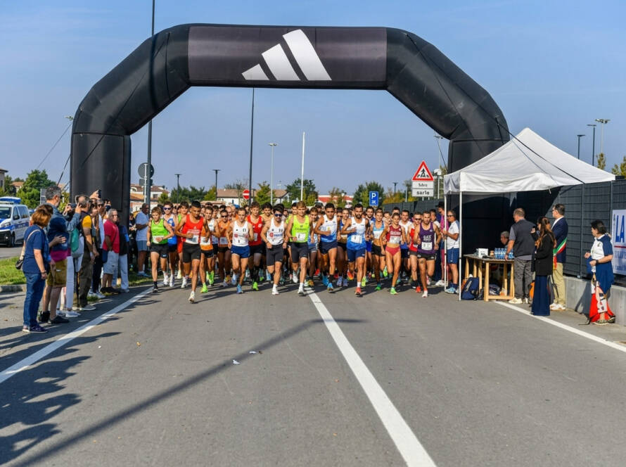 Gruppo numeroso di podisti in tenuta sportiva al momento della partenza della gara Nova Run, mentre attraversano la linea sotto un grande arco gonfiabile nero targato Adidas. Sulla destra sono presenti un gazebo bianco, transenne con spettatori e il cartellone pubblicitario La Rocca.
