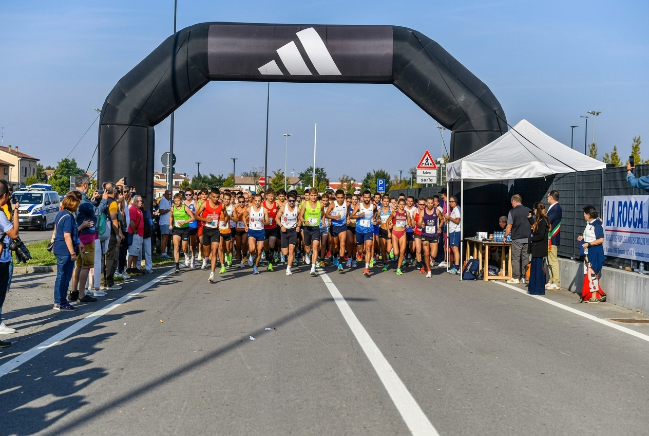Gruppo numeroso di podisti in tenuta sportiva al momento della partenza della gara Nova Run, mentre attraversano la linea sotto un grande arco gonfiabile nero targato Adidas. Sulla destra sono presenti un gazebo bianco, transenne con spettatori e il cartellone pubblicitario La Rocca.